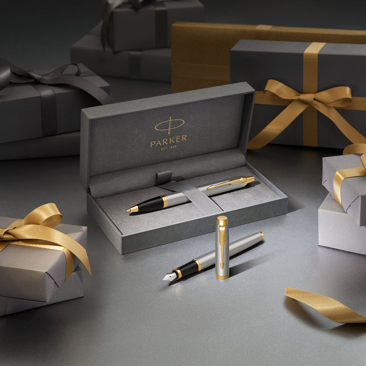 Pióro wieczne Parker IM Brushed Metal GT w pudełku prezentowym z długopisem, otoczone eleganckimi szaro-złotymi paczkami