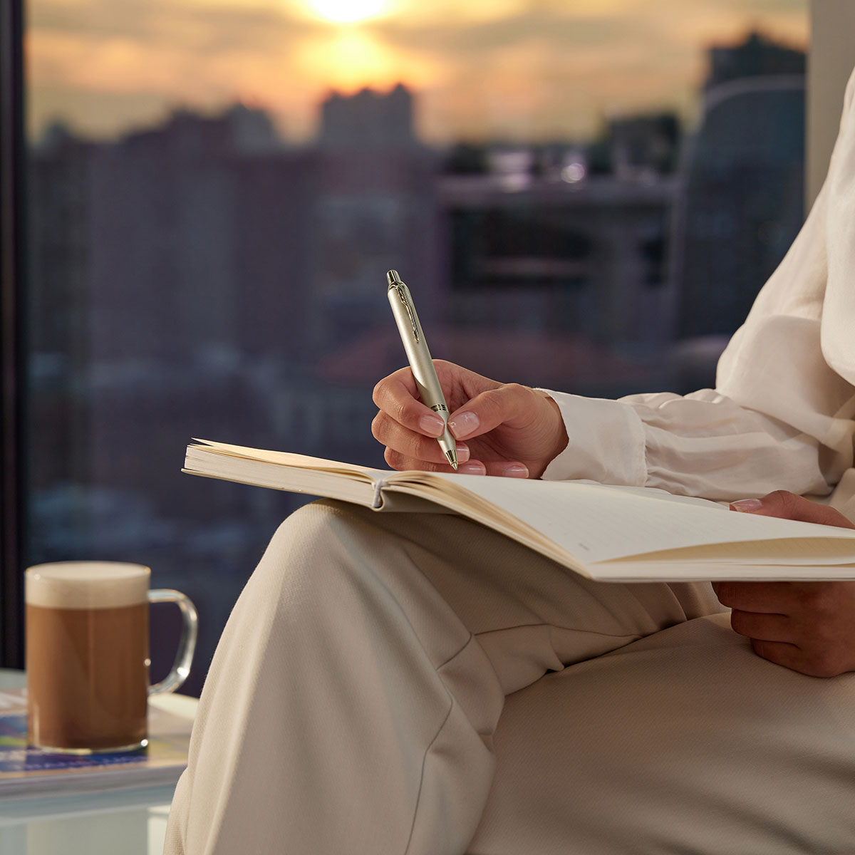 Długopis Parker IM Monochrome Champagne w dłoni kobiety piszącej w notatniku przy biurku z laptopem i rośliną