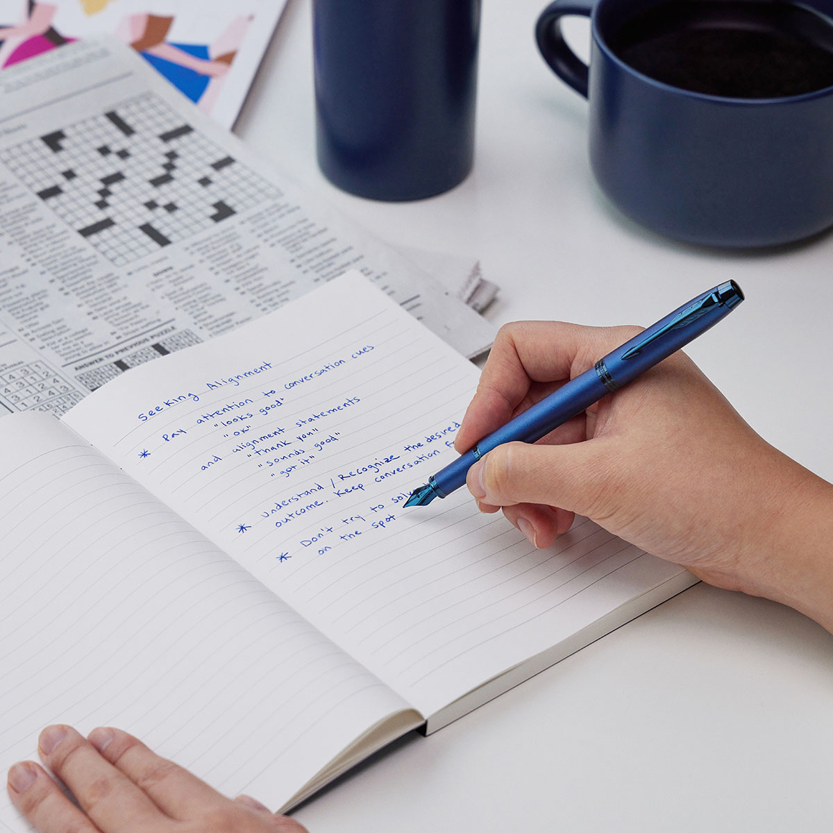 Pióro Parker IM Professionals Monochrome Blue (2172966/2) używane do pisania w notesie, obok granatowego kubka i gazety.