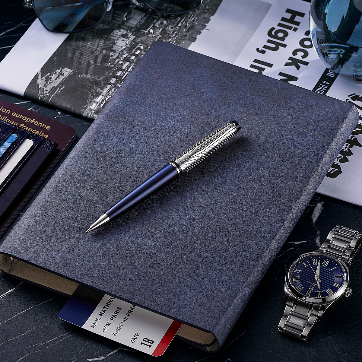 długopis Waterman Expert Deluxe Metalic Niebieski na granatowym notesie obok zegarka i biletu lotniczego