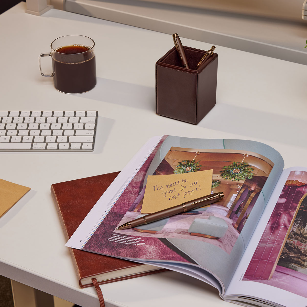 Pióro kulkowe Parker IM Monochrome Burgundy CT leżące na otwartym magazynie, z notatką na żółtej karteczce i filiżanką kawy w tle – scena z biurka.