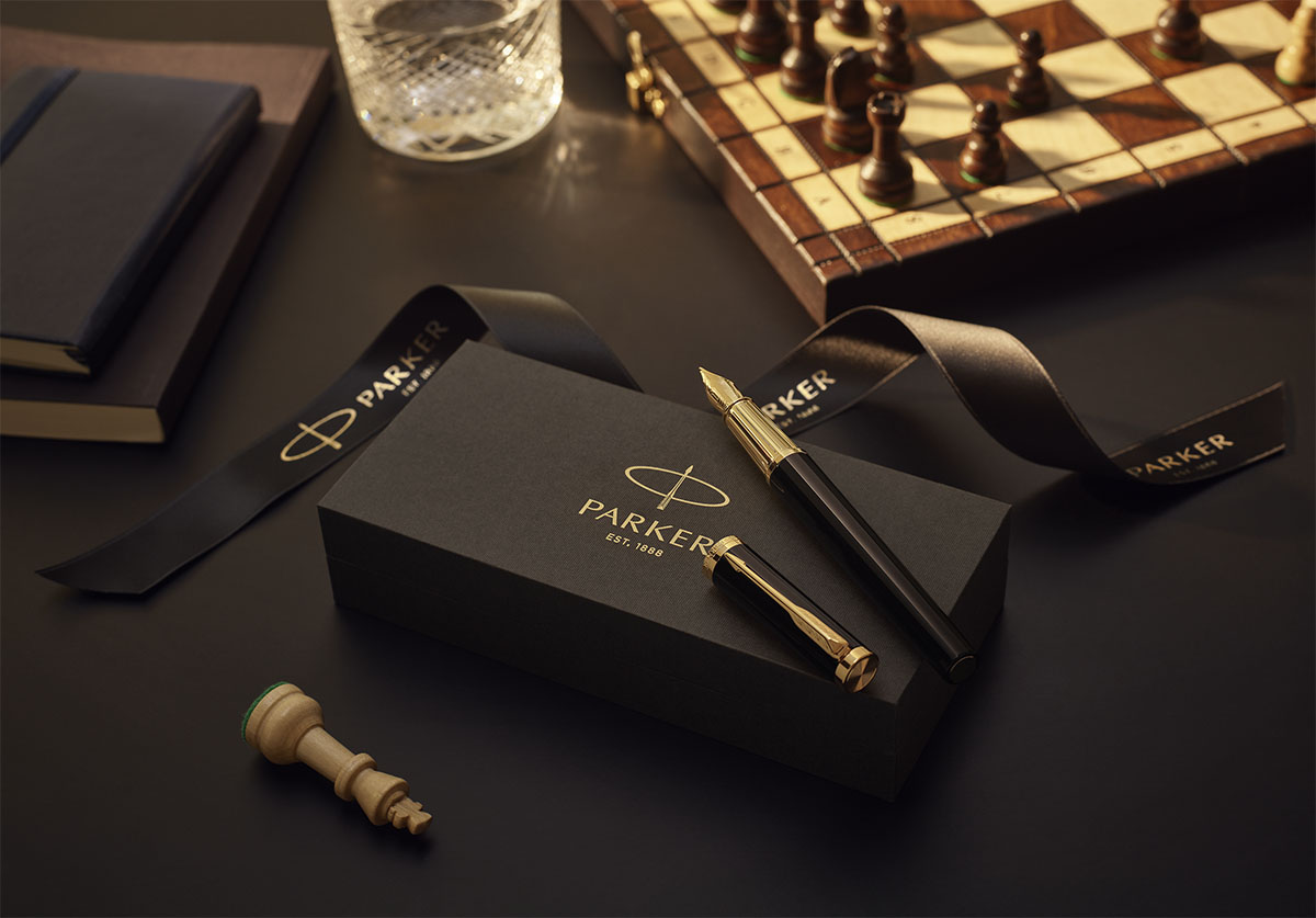Luksusowe pióro wieczne Parker z wykończeniem GT na eleganckim czarnym pudełku prezentowym – w tle szachownica, szkło do whisky i notatniki.