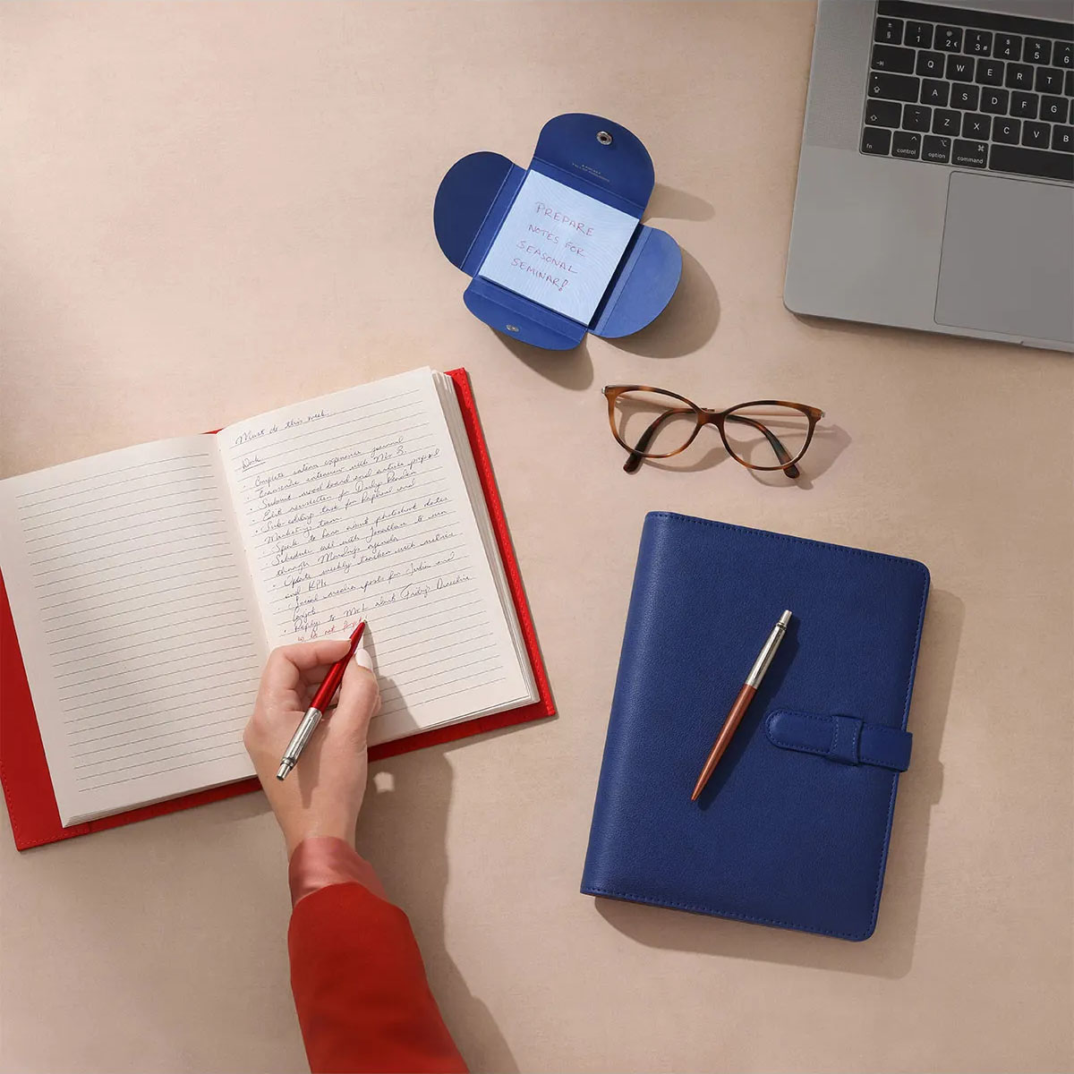 Osoba pisze w notesie czerwonym długopisem, obok laptop, okulary i niebieski organizer