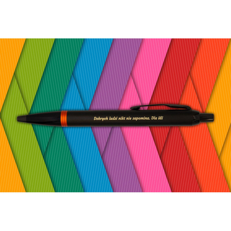 Czarny długopis Parker IM Professionals Flame Orange z pomarańczowym pierścieniem i grawerem „Dobrych ludzi nikt nie zapomina. Dla Lili” na tle kolorowych pasków.