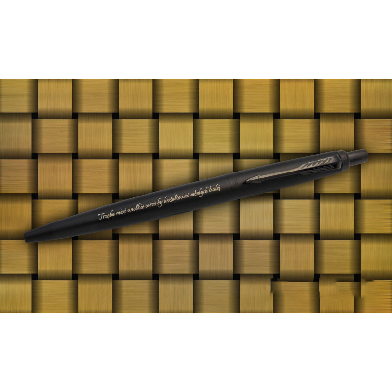 Czarny długopis Parker Jotter XL Monochrome z grawerem "Trzeba mieć wielkie serce by kształtować młodych ludzi" na tle złotej plecionki.