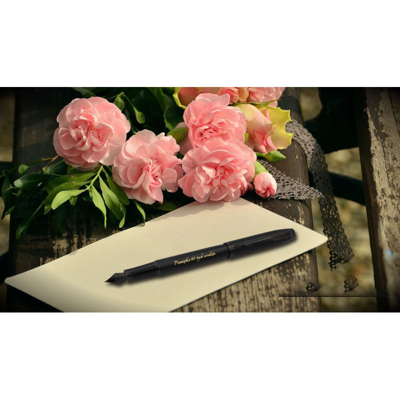 Czarne pióro wieczne Parker IM Achromatic z grawerem „Pamiątka do życia w radości” w złotym kolorze, ułożone na białej kartce, obok bukietu różowych kwiatów