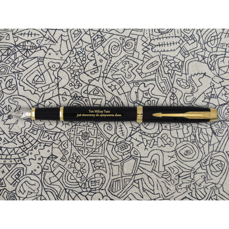 Czarne pióro wieczne Parker IM GT z pozłacanymi detalami, w tym klipsem w kształcie strzały i pierścieniami, z grawerem w kolorze złotym o treści „Ten Wielcy Tusz jest stworzony do opisywania dusz”. Pióro leży na tle z fantazyjnymi, ręcznie rysowanymi cza
