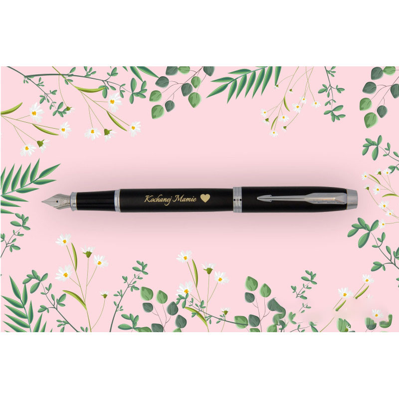 Czarne, matowe pióro wieczne Parker IM CT ze srebrnymi wykończeniami i charakterystycznym klipsem w kształcie strzały, ozdobione złotym grawerem „Kochanej Mamie” z symbolem serca. Pióro umieszczone na pastelowym, różowym tle z ilustracjami zielonych liści