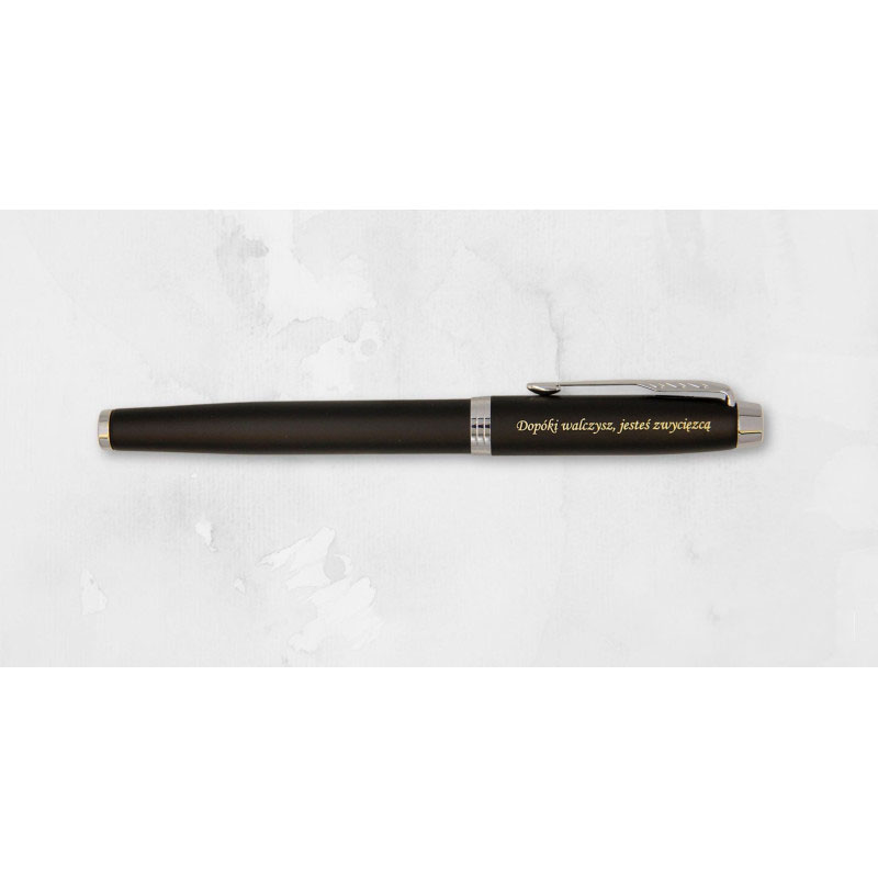 Czarne, matowe pióro wieczne Parker IM CT z chromowanymi wykończeniami i charakterystycznym klipsem w kształcie strzały. Na korpusie znajduje się złoty grawer z napisem „Dopóki walczysz, jesteś zwycięzcą”. Produkt sfotografowany na jasnoszarym tle przypom