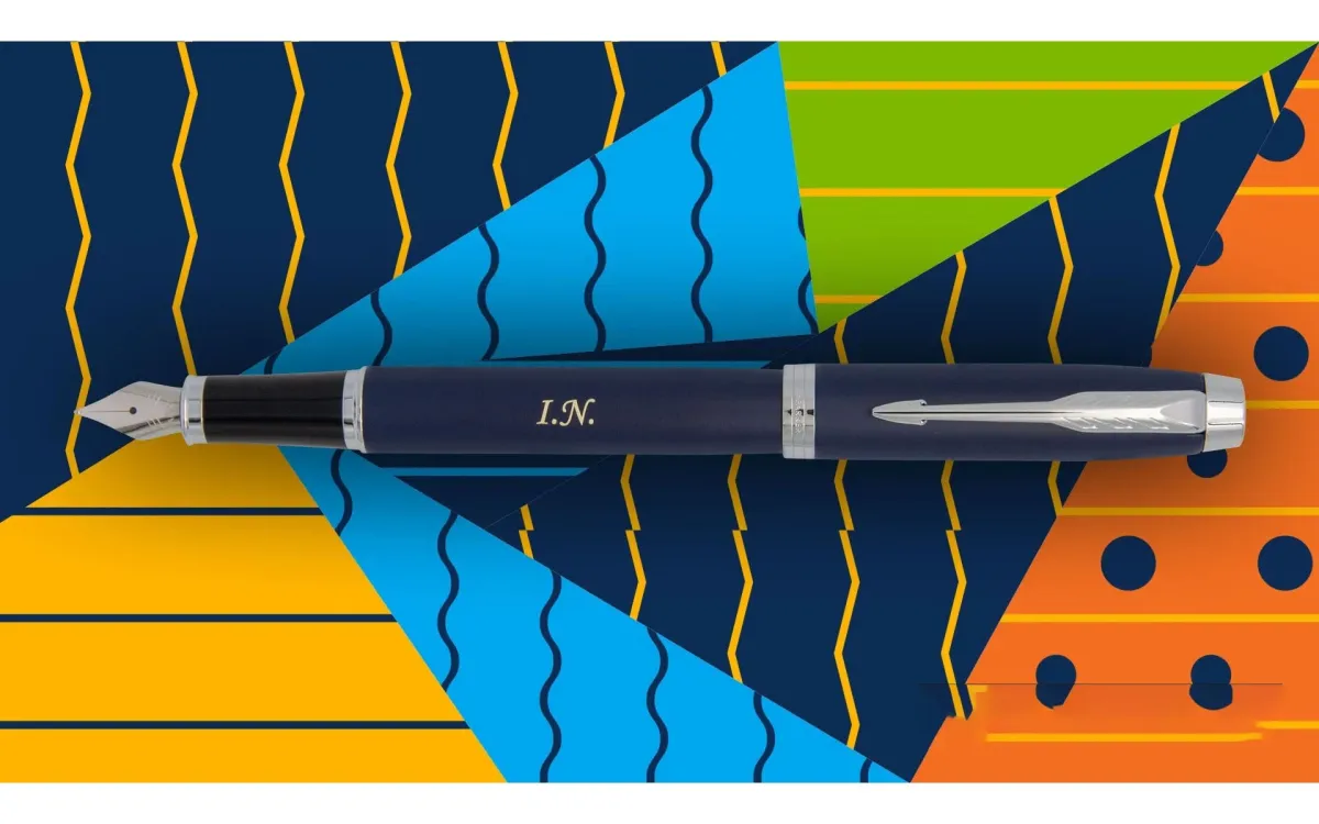 Pi&oacute;ro wieczne Parker IM Niebieskie CT [1931647] &ndash; eleganckie granatowe pi&oacute;ro z wykończeniami w kolorze chromu (CT &ndash; Chrome Trim), grawer: &bdquo;I.N.&rdquo;. Umieszczone na tle kolorowej, geometrycznej grafiki w odcieniach ż&oacute;łci, błękitu, zieleni i pomarańczu.