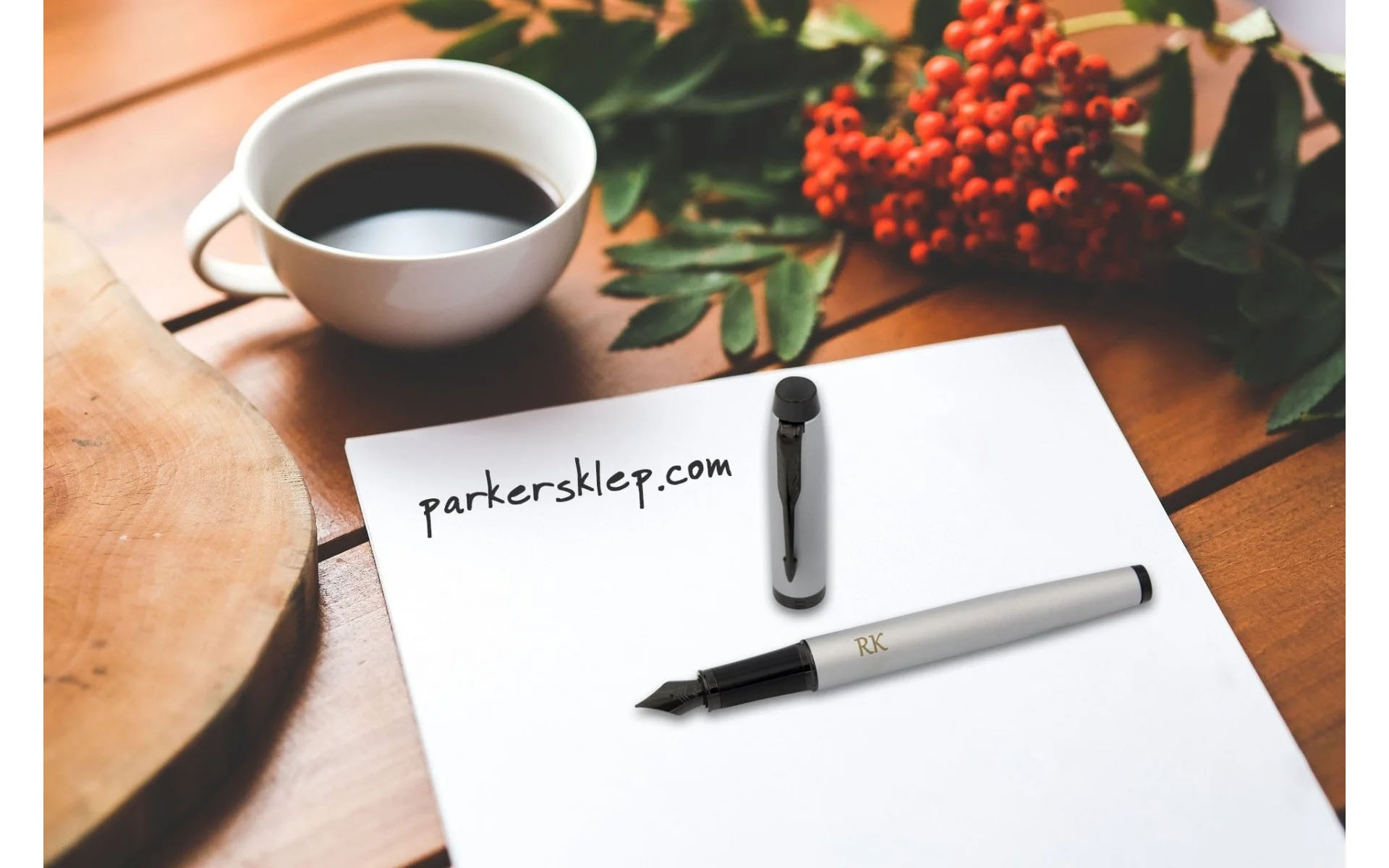 Pióro wieczne Parker IM Achromatic Srebrne [2127619] – eleganckie pióro w matowym srebrnym kolorze z czarnymi wykończeniami. Na korpusie grawer „RK”. Ujęcie na stole z filiżanką kawy, czerwonymi owocami jarzębiny i kartką papieru z napisem „parkersklep.co