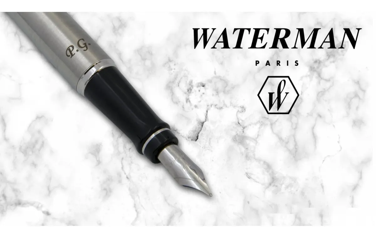 Pi&oacute;ro wieczne Waterman Expert stalowe CT [S0952040] z inicjałami &bdquo;P.G.&rdquo; wygrawerowanymi na korpusie, zaprezentowane na tle białego marmuru obok logo marki Waterman Paris.