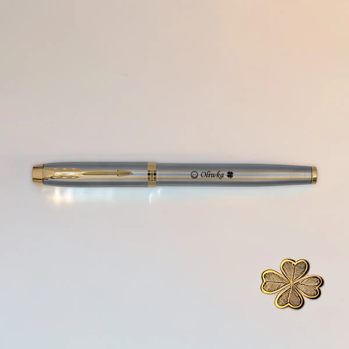 Pi&oacute;ro wieczne Parker IM Brushed Metal GT (1931649) z grawerem &bdquo;Oliwka&rdquo; i symbolem koniczynki, sfotografowany na jasnym tle obok złotej czterolistnej koniczynki.