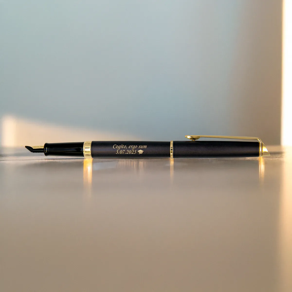 Pi&oacute;ro wieczne Waterman Hemisphere Lava Black GT (2217939) z grawerem &bdquo;Cogito, ergo sum &ndash; 3.07.2025&rdquo; na stole w ciepłym świetle zachodzącego słońca, z delikatnym złotym refleksem.