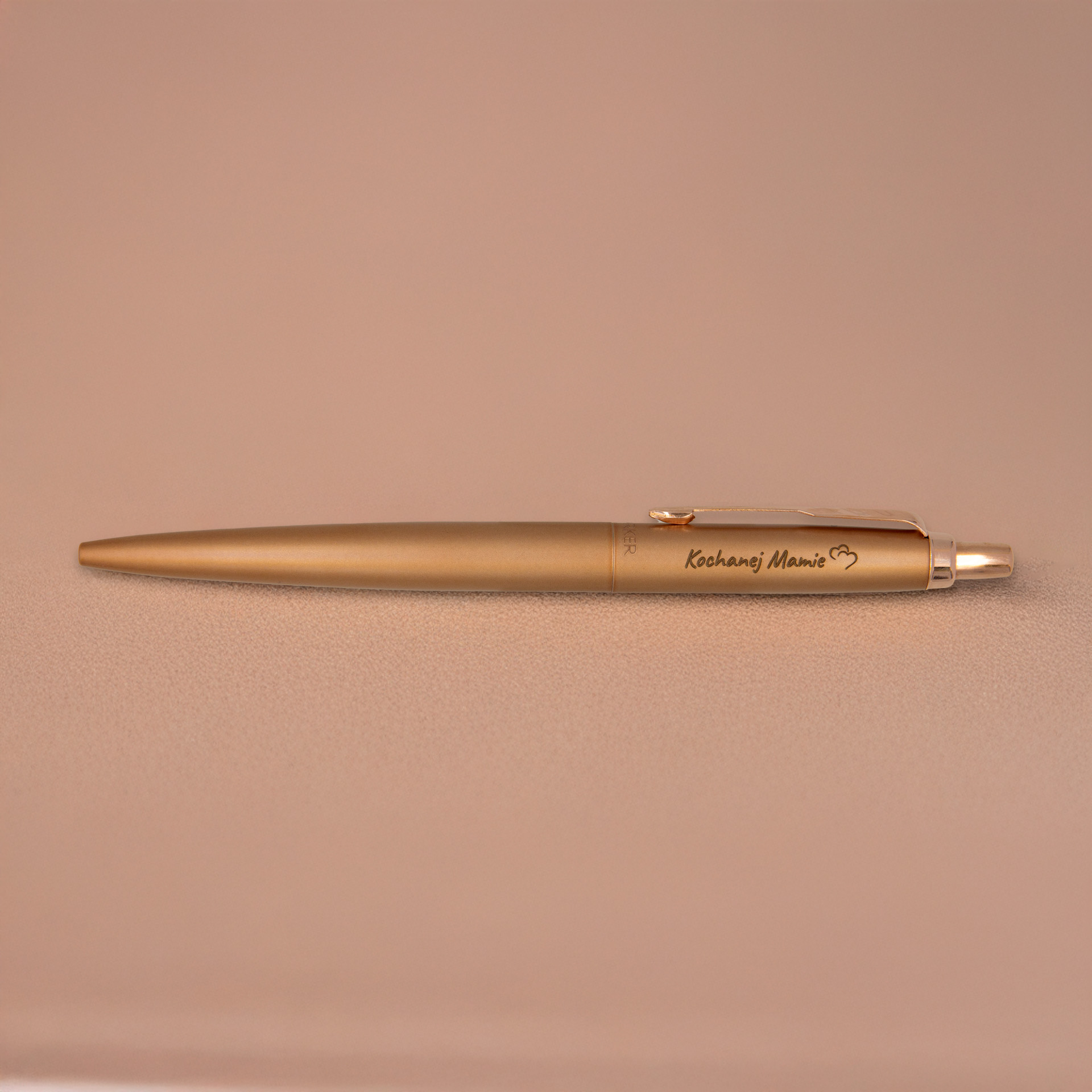 Długopis Parker Jotter XL Monochrome Pink Gold (2122755) z grawerem „Kochanej Mamie” i sercem – elegancki upominek w ciepłym odcieniu brązowego złota, sfotografowany na beżowym tle.