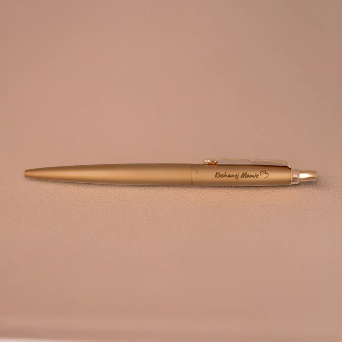 Długopis Parker Jotter XL Monochrome Pink Gold (2122755) z grawerem „Kochanej Mamie” i sercem – elegancki upominek w ciepłym odcieniu brązowego złota, sfotografowany na beżowym tle.