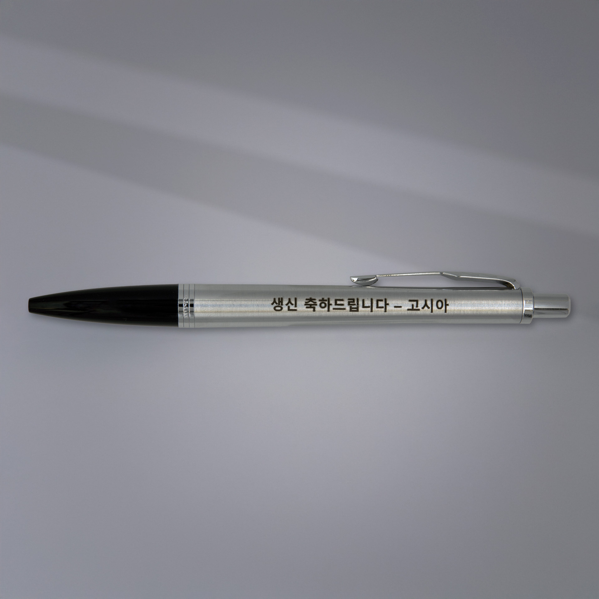 Długopis Parker stalowy z czarnym wykończeniem i grawerem w języku koreańskim „생신 축하드립니다 – 고시아”, eleganckie ujęcie na tle z delikatnym cieniem światła.