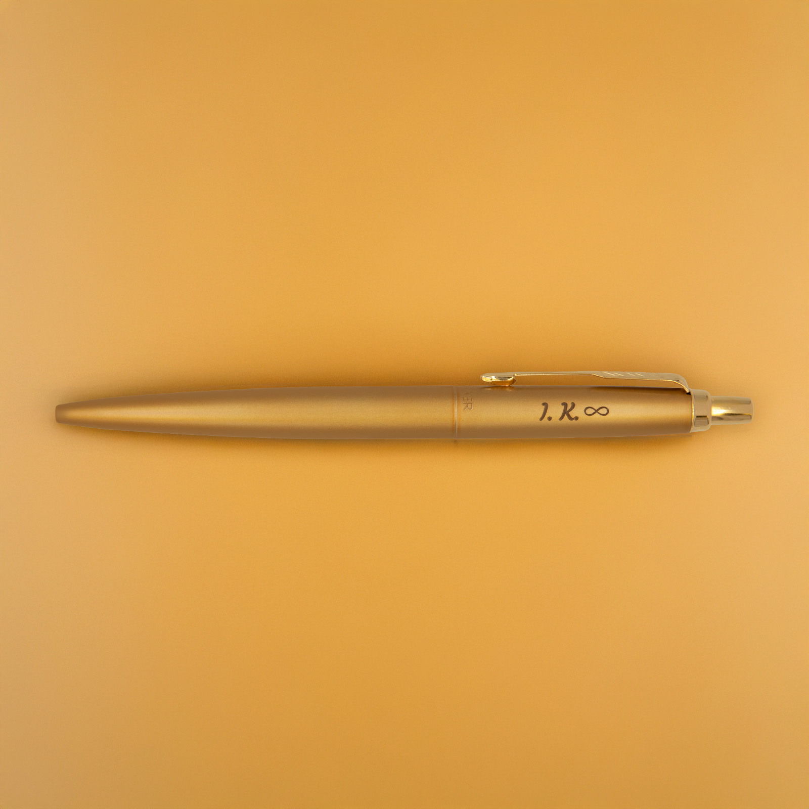 Długopis Parker Jotter XL Monochrome Gold z grawerem „I.K. ∞”, sfotografowany na złotym tle w ciepłym świetle, podkreślającym jego jednolity kolor i połysk.