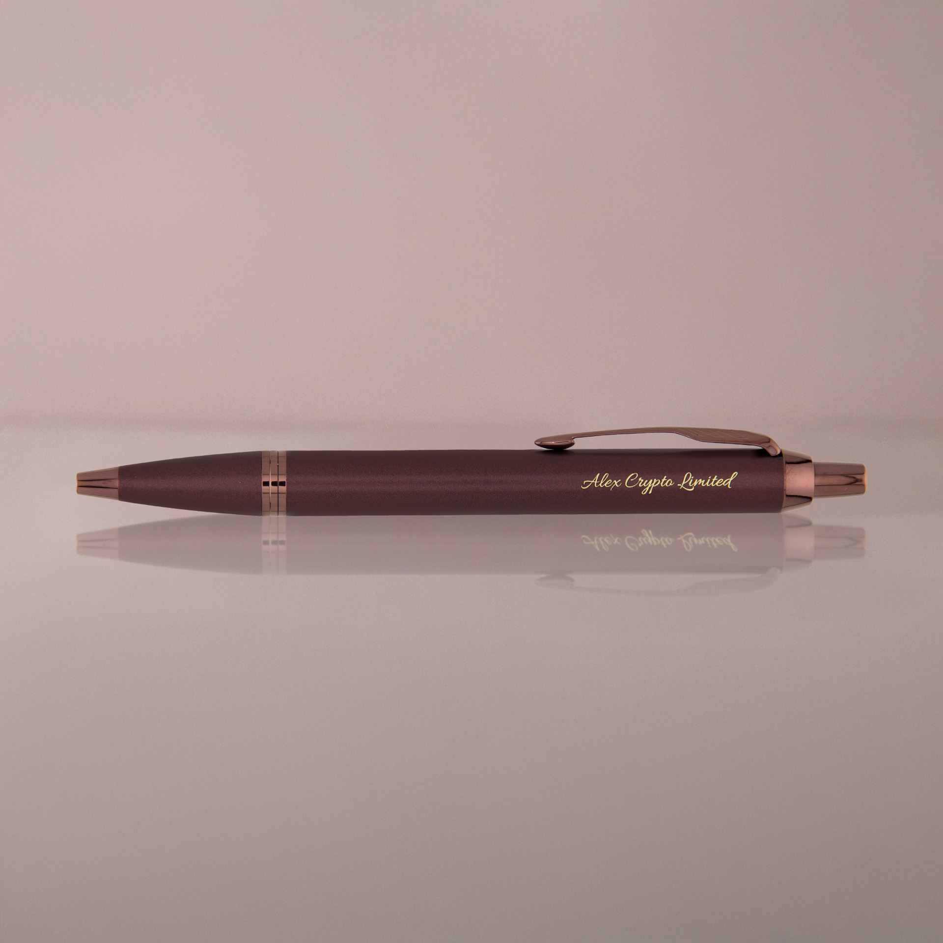 Brązowy długopis z miedzianymi wykończeniami i grawerem „Alex Crypto Limited” na różowo-beżowym tle.