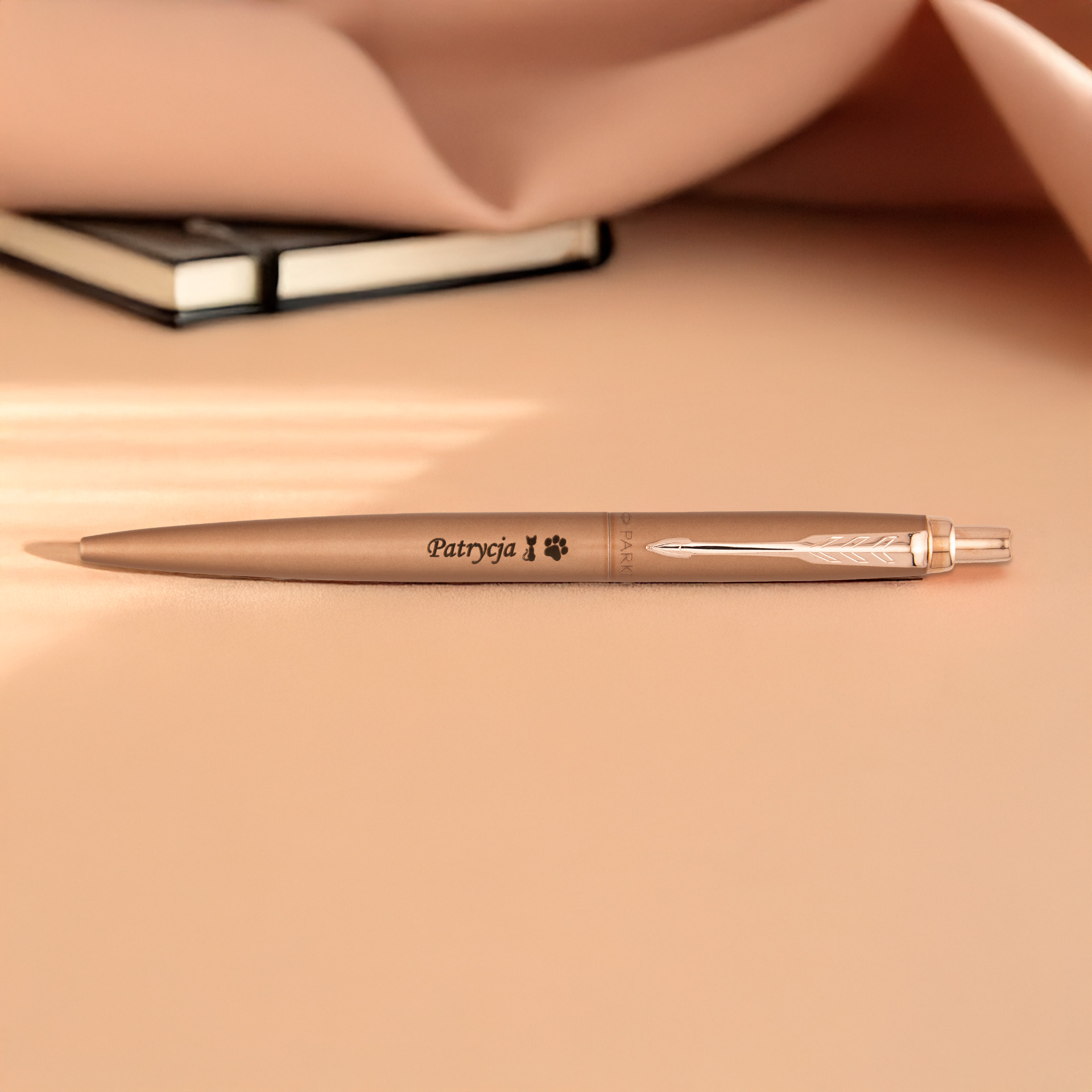 Matowy długopis Parker Jotter XL Pink Gold z grawerem „Patrycja” leży na beżowym tle obok notesu.