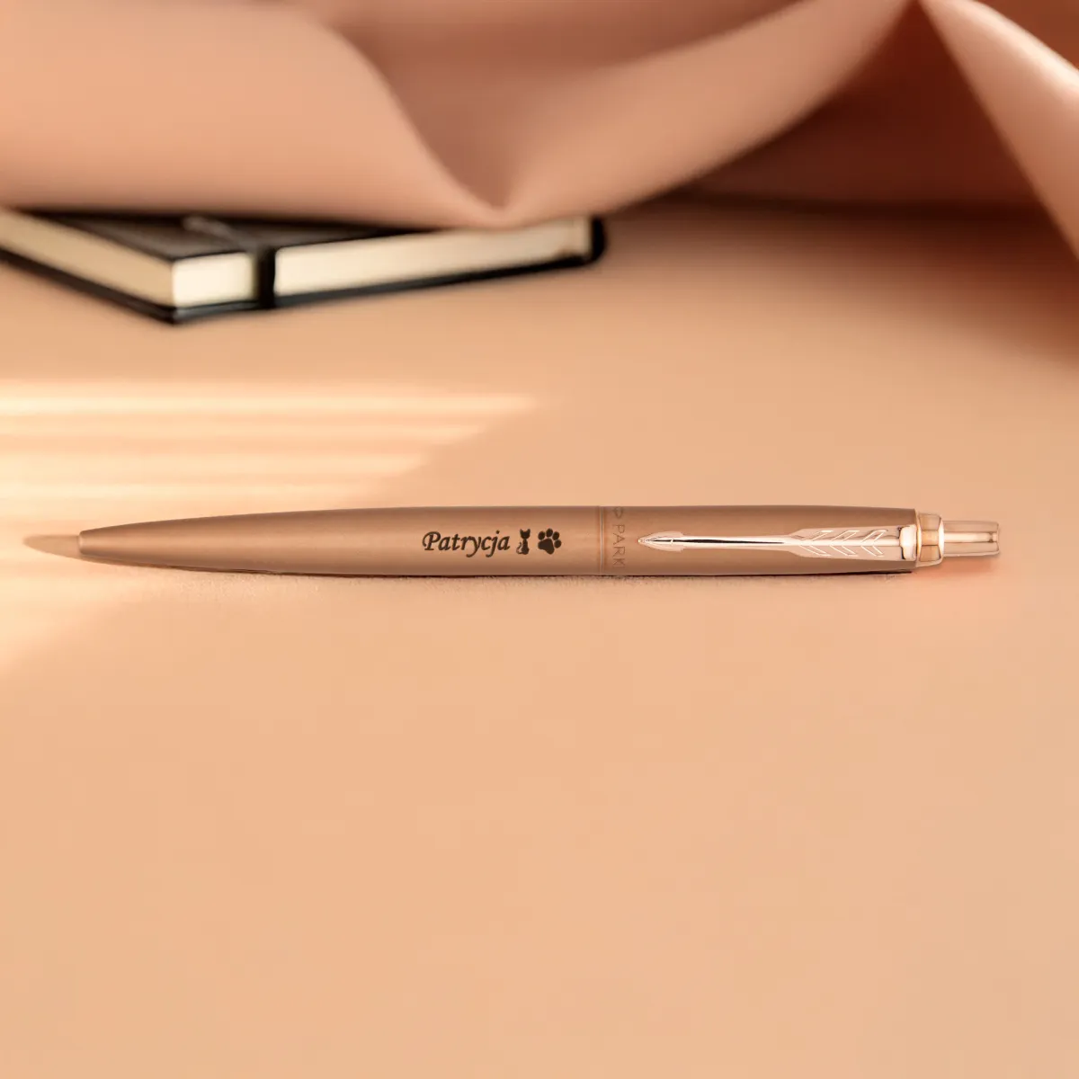 Matowy długopis Parker Jotter XL Pink Gold z grawerem „Patrycja” leży na beżowym tle obok notesu.