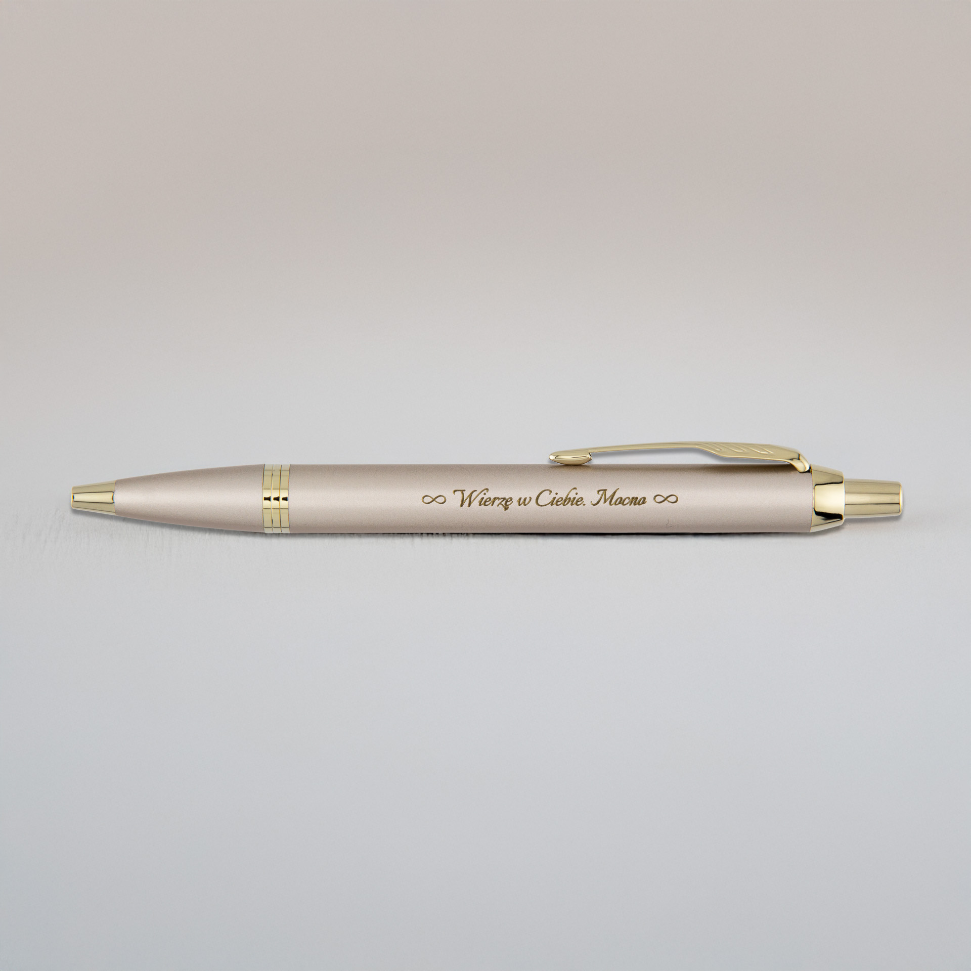 Jasny, złotawy długopis z grawerem „Wierzę w Ciebie. Mama” leży na neutralnym tle.