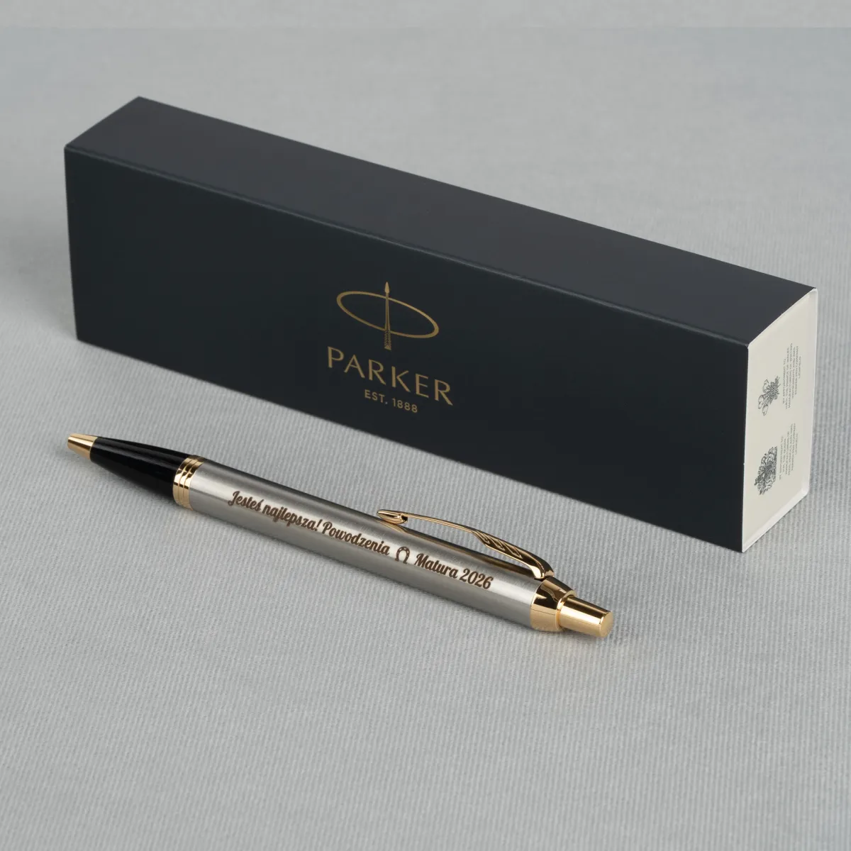 Długopis Parker IM Brushed Metal GT z grawerem „Jesteś najlepsza! Powodzenia Matura 2026” na jasnym tle z pudełkiem Parker w tle