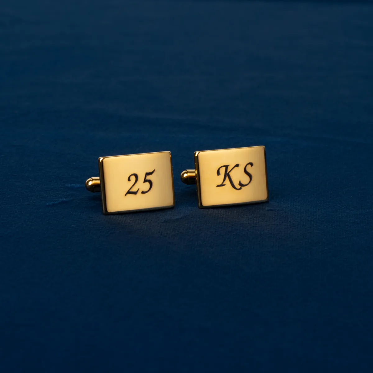 Złote stalowe spinki do koszuli z grawerem „25” oraz inicjałami „KS” na granatowym tle.