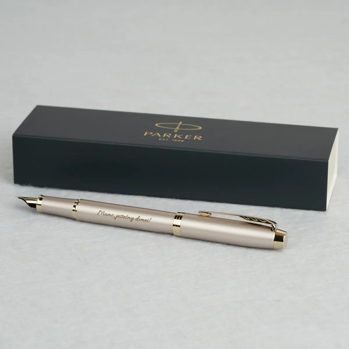 Pióro wieczne Parker IM Professionals Monochrome Champagne z grawerem „Mamo, jesteśmy dumni!”, ułożone przed eleganckim etui Parker.