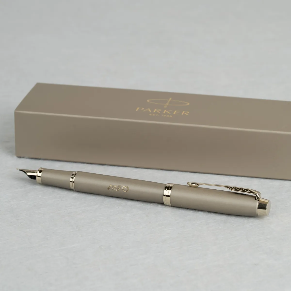 Pióro wieczne Parker IM Rituals Sandstone w piaskowym, matowym wykończeniu z grawerem inicjałów „MF” i symbolem serca, prezentowane na tle eleganckiego firmowego etui Parker
