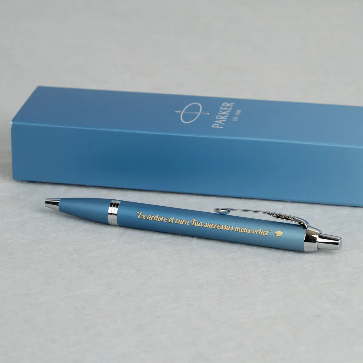 Długopis Parker IM Rituals Turquoise Blue CT w matowym turkusowym wykończeniu z grawerowaną sentencją oraz symbolem biretu, prezentowany na tle eleganckiego niebieskiego etui Parker